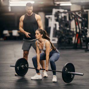 A strong sportswoman is lifting weights in a gym while her instructor is helping her.