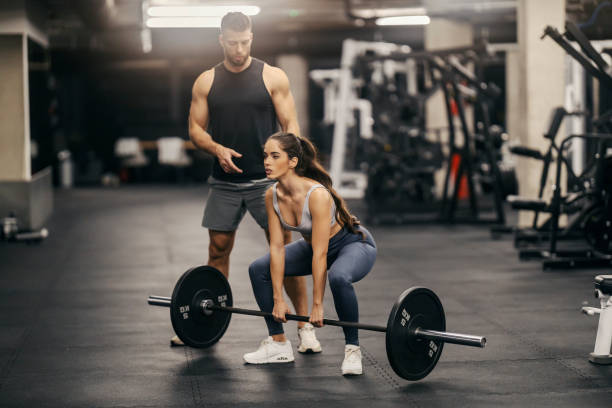 A strong sportswoman is lifting weights in a gym while her instructor is helping her.